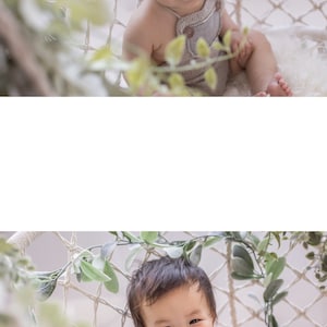 May include: A baby wearing a light brown overall with a white background. The baby is sitting in a white hammock with green leaves hanging from it. The baby is smiling and looking at the camera.