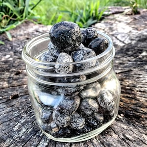 Pode incluir: Um frasco de vidro cheio de pedras de obsidiana negra polidas. As pedras são lisas e têm um aspecto brilhante, quase húmido.