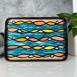 May include: A wooden tray with a black frame and a painted design of wavy lines in shades of blue, orange, and yellow. The design resembles water with waves.