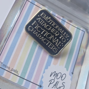 May include: A black enamel pin with gold edging, inscribed with "Emotionally Attached to Fictional Characters." The pin is presented on a pastel striped card, packaged in a clear plastic bag. The Moo Pals logo is also visible.