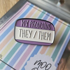 May include: Purple enamel pin with white lettering that reads "My Pronouns They/Them".