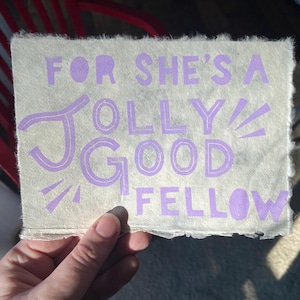 May include: A rectangular, off-white paper with the text "FOR SHE'S A JOLLY GOOD FELLOW" in lavender. The paper has a rough, textured edge. The image is held by a hand.