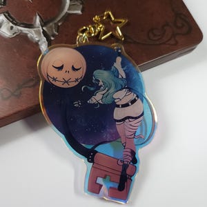 Könnte beinhalten: Ein klarer Acryl-Schlüsselanhänger mit einer Cartoon-Illustration eines Charakters mit blauen Haaren und mumienartigen Bandagen, vor einem Sternenhimmel und einem Jack-Skellington-ähnlichen Mond. Der Schlüsselanhänger hat einen goldfarbenen sternförmigen Verschluss.