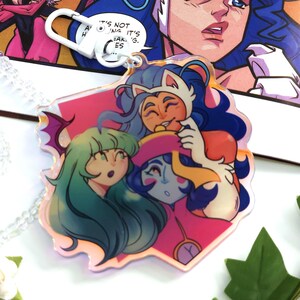 May include: A colorful acrylic charm featuring three cartoon characters with long hair and unique outfits. The charm has a white clasp and a pink, iridescent background. The text "IT'S NOT WORKING. IT'S FAKING. ES" is visible in the background.