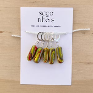 May include: Set of five colorful glass stitch markers with silver metal loops. The markers are yellow, red, and green with a marbled pattern. The card reads "sego fibers" and "Progress Keepers & Stitch Markers".