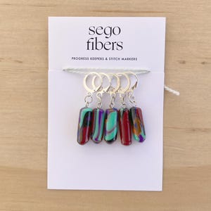 May include: Set of five colorful glass stitch markers with silver metal loops. The markers are red, purple, and green with a marbled pattern. The card reads "sego fibers" and "Progress Keepers & Stitch Markers".