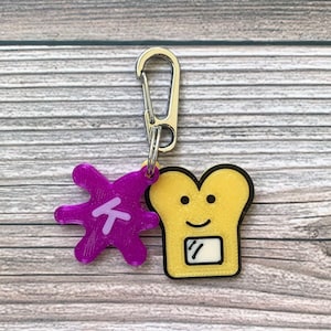 Puede incluir: Un llavero morado con una letra T y un amuleto de pan de dibujos animados amarillo con una cara sonriente.