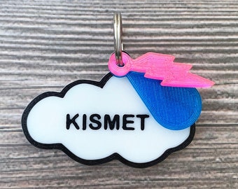 May include: White cloud-shaped keychain with black outline and the word "KISMET" in black lettering. A blue and pink lightning bolt charm is attached to the keychain.