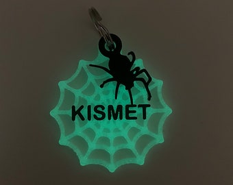 May include: A glow-in-the-dark spider web keychain with a black spider silhouette and the word "KISMET" written in black.