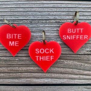 Puede incluir: Tres llaveros de corazón rojo con texto blanco. Los llaveros dicen "Bite Me", "Sock Thief" y "Butt Sniffer".