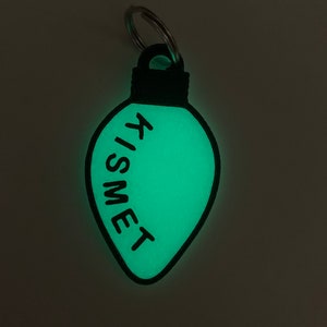 Op de afbeelding: Een groene, in het donker lichtgevende gloeilamp sleutelhanger met het woord "KISMET" erop.