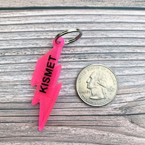 May include: A pink lightning bolt keychain with the word "Kismet" printed on it. The keychain is attached to a silver key ring.