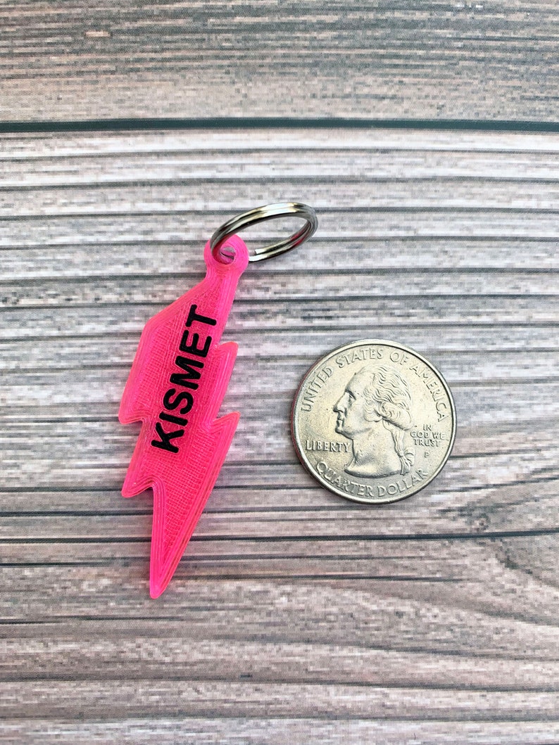 May include: A pink lightning bolt keychain with the word "Kismet" printed on it. The keychain is attached to a silver key ring.