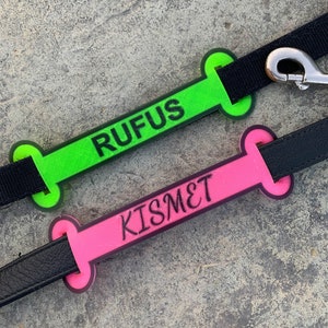 May include: Two dog leash tags, one green and one pink, shaped like bones. The green tag says "RUFUS" and the pink tag says "KISMET".