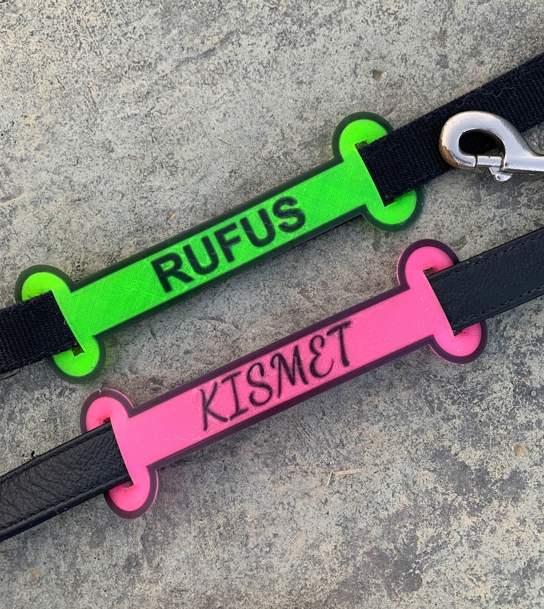 May include: Two dog leash tags, one green and one pink, shaped like bones. The green tag says "RUFUS" and the pink tag says "KISMET".
