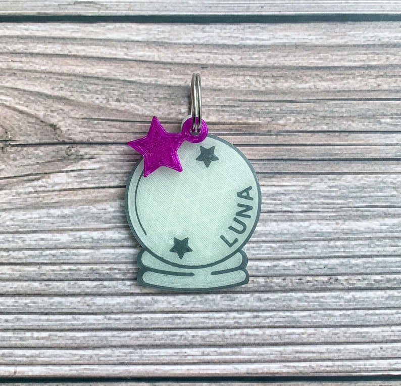 May include: A white and gray 3D printed charm shaped like a crystal ball with a silver ring at the top. The charm has a purple star on top and the word "LUNA" written on the side.