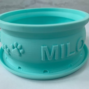 Puede incluir: Un cuenco para mascotas de plástico azul claro con un borde elevado y una base antideslizante. El cuenco tiene un diseño de huella de pata y el nombre "MILO" impreso en el lateral.