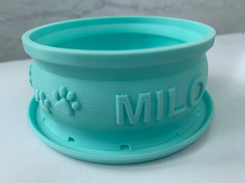 Peut inclure: Un bol pour animal de compagnie en plastique bleu clair avec un bord sur&eacute;lev&eacute; et une base antid&eacute;rapante. Le bol est d&eacute;cor&eacute; d'une empreinte de patte et le nom "MILO" est imprim&eacute; sur le c&ocirc;t&eacute;.