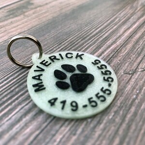 May include: A white glow-in-the-dark dog tag with a black paw print and the text "Maverick 419-555-5555".