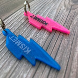 May include: Two lightning bolt shaped keychains, one pink and one blue, with the word "Kismet" printed on each.