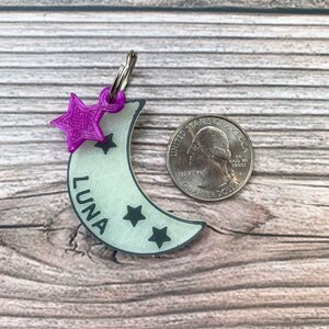 May include: A white crescent moon-shaped dog tag with the name "LUNA" and three black stars. A purple star charm is attached to the top of the tag.
