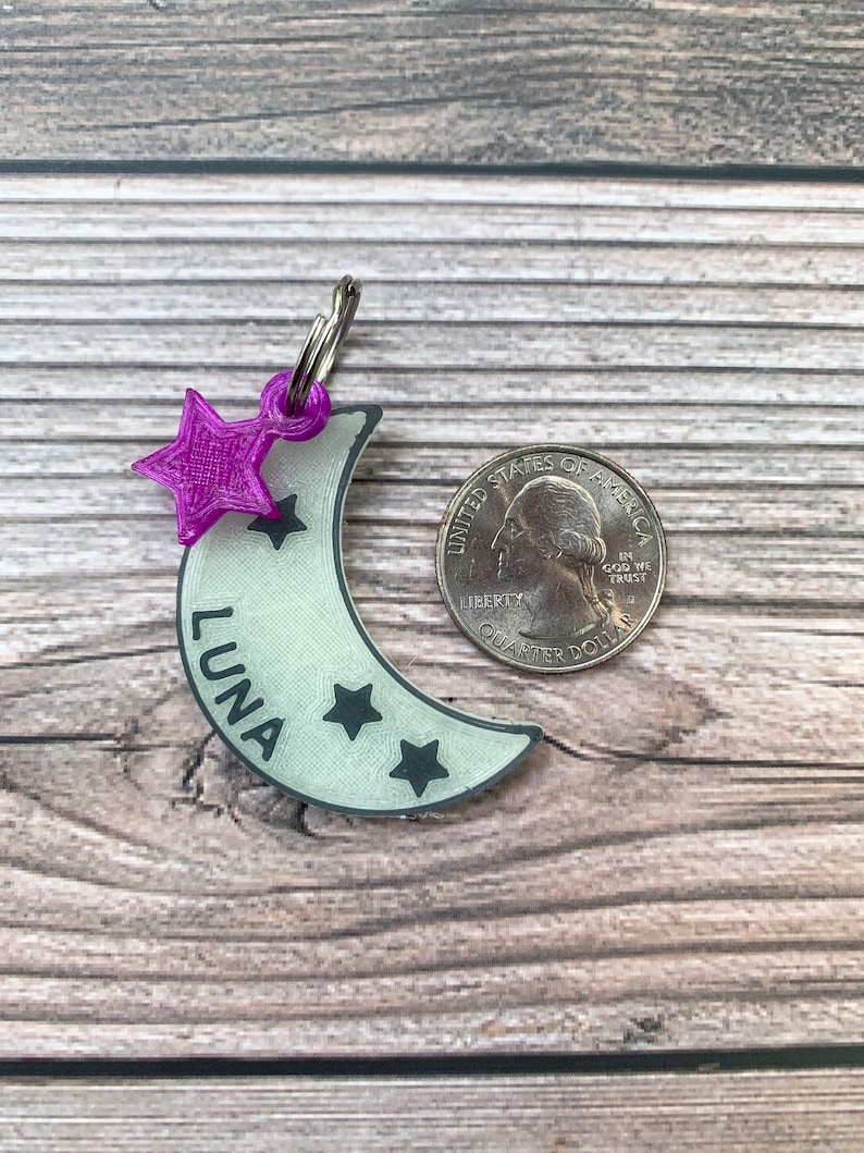 May include: A white crescent moon-shaped dog tag with the name "LUNA" and three black stars. A purple star charm is attached to the top of the tag.