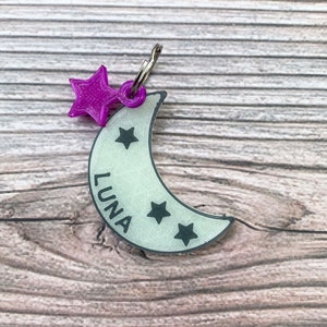 May include: A white crescent moon-shaped dog tag with the name "LUNA" printed on it. The tag has a purple star charm attached to it and four black stars on the moon.