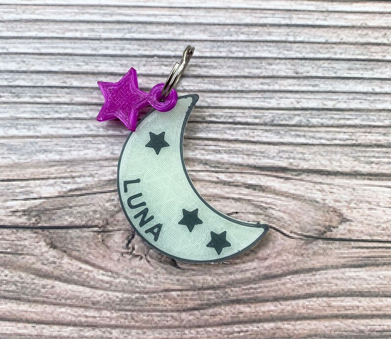 May include: A white crescent moon-shaped dog tag with the name "LUNA" printed on it. The tag has a purple star charm attached to it and four black stars on the moon.