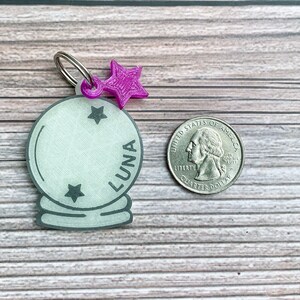 May include: A white plastic charm in the shape of a crystal ball with a silver ring attached. The charm has three black stars and the word "LUNA" written on it. A purple star charm is attached to the ring.