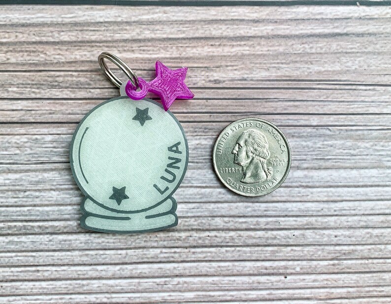 May include: A white plastic charm in the shape of a crystal ball with a silver ring attached. The charm has three black stars and the word "LUNA" written on it. A purple star charm is attached to the ring.