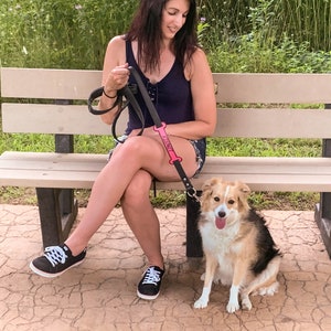May include: A woman wearing a black tank top and jeans sits on a bench with a dog on a leash. The leash has a pink bone-shaped tag that says "Paws". The dog is a brown and white mix with a long tail.