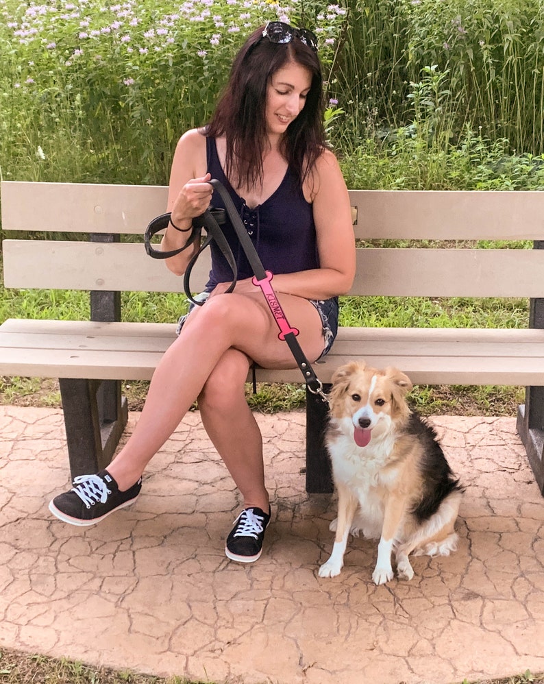 May include: A woman wearing a black tank top and jeans sits on a bench with a dog on a leash. The leash has a pink bone-shaped tag that says "Paws". The dog is a brown and white mix with a long tail.
