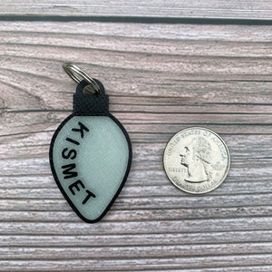 Op de afbeelding: Een witte lichtbollen ornament sleutelhanger met het woord "TISMET" erop geschreven in het zwart. De ornament is ongeveer zo groot als een Nederlandse 2 euro munt.