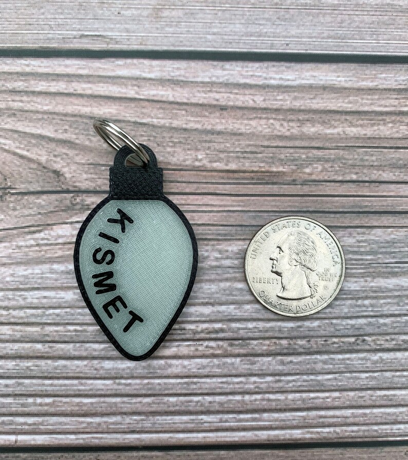 Op de afbeelding: Een witte lichtbollen ornament sleutelhanger met het woord "TISMET" erop geschreven in het zwart. De ornament is ongeveer zo groot als een Nederlandse 2 euro munt.