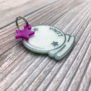 May include: A white plastic charm shaped like a crystal ball with the word "LUNA" written on it. A purple star charm is attached to a silver ring.