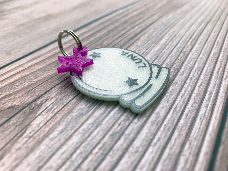 May include: A white plastic charm shaped like a crystal ball with the word "LUNA" written on it. A purple star charm is attached to a silver ring.