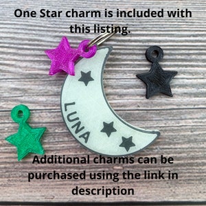 May include: A white crescent moon charm with the word "LUNA" inscribed on it. The charm has three small stars on it and is attached to a silver ring. A pink star charm, a green star charm, and a black star charm are also shown.