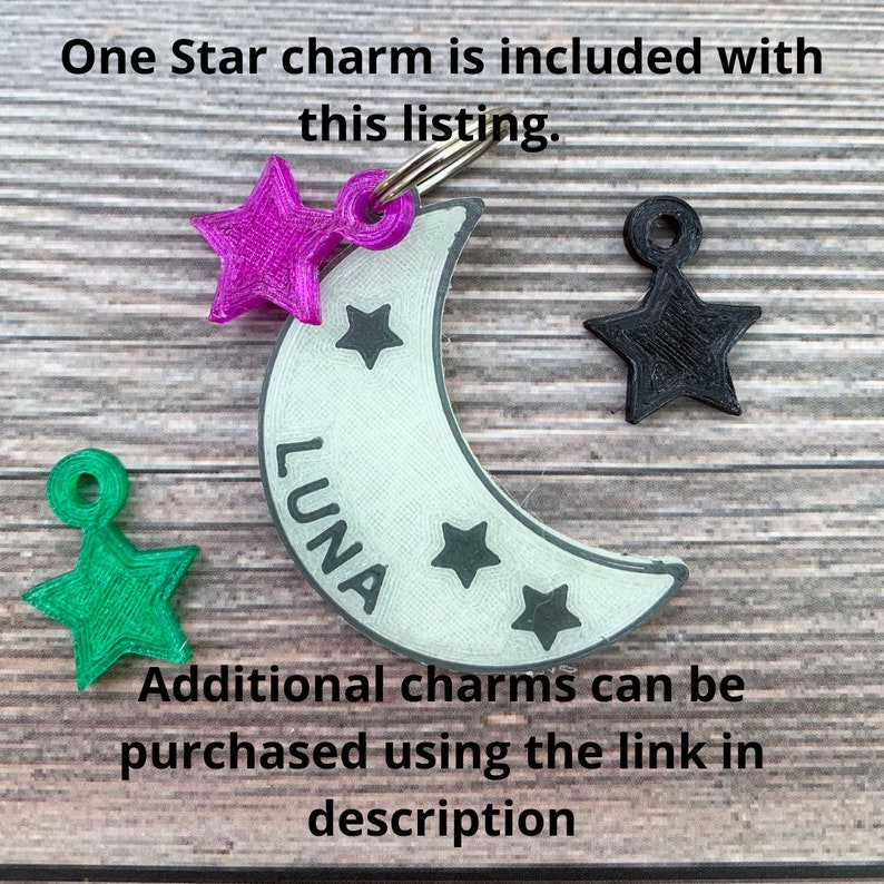 May include: A white crescent moon charm with the word "LUNA" inscribed on it. The charm has three small stars on it and is attached to a silver ring. A pink star charm, a green star charm, and a black star charm are also shown.