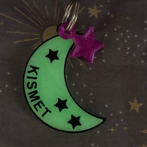 May include: A green glow-in-the-dark crescent moon keychain with the word "KISMET" written vertically on the side. The keychain has three black stars on the moon and a purple star charm attached to a silver key ring.