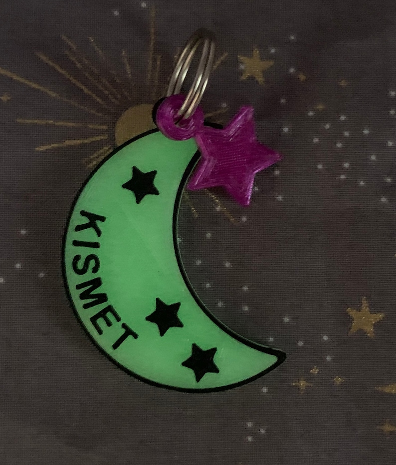 May include: A green glow-in-the-dark crescent moon keychain with the word "KISMET" written vertically on the side. The keychain has three black stars on the moon and a purple star charm attached to a silver key ring.