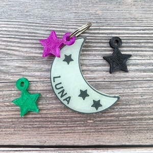 May include: A white crescent moon-shaped dog tag with the name "LUNA" printed on it. The tag has three black stars and is attached to a silver key ring. There are three star-shaped charms in pink, green, and black.