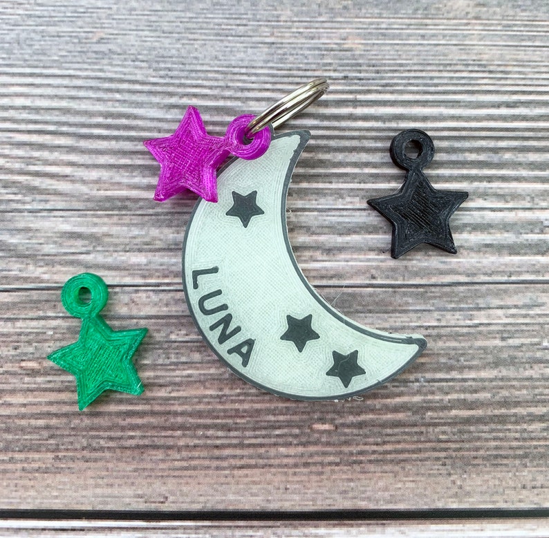 May include: A white crescent moon-shaped dog tag with the name "LUNA" printed on it. The tag has three black stars and is attached to a silver key ring. There are three star-shaped charms in pink, green, and black.