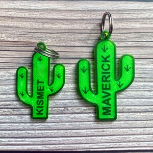 May include: Two green cactus-shaped keychains with the names "Maverick" and "Kismet" printed on them. The keychains are made of a 3D printed material and have a textured surface.