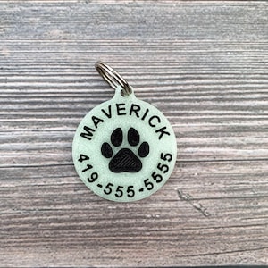 May include: A round, light green pet ID tag with a black paw print and the text "MAVERICK 419-555-5555".