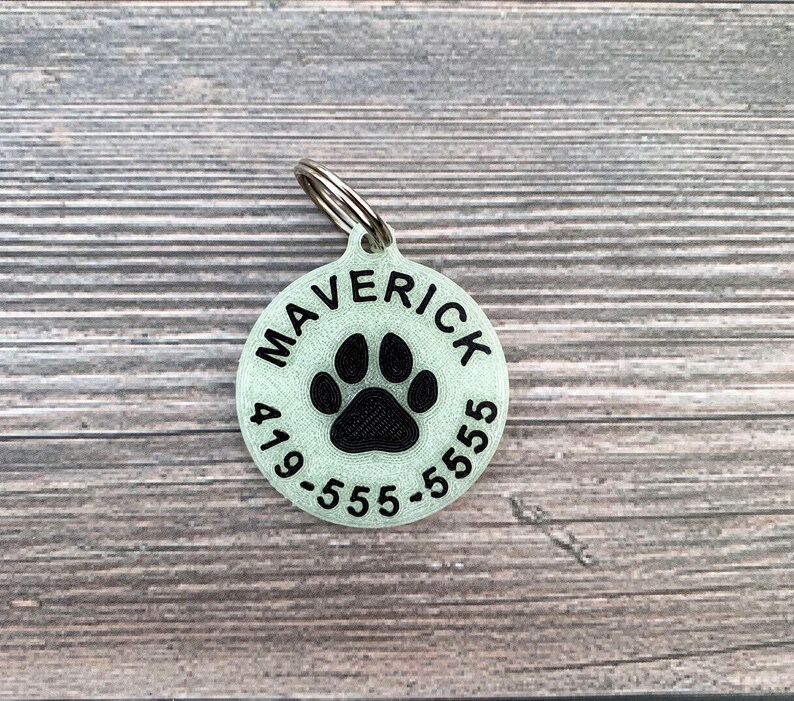 May include: A round, light green pet ID tag with a black paw print and the text "MAVERICK 419-555-5555".