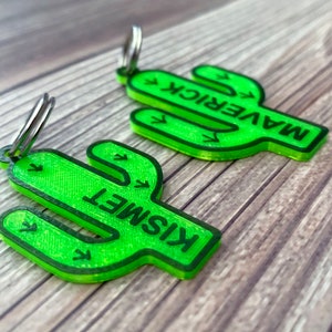 May include: Two green cactus-shaped keychains with silver key rings. The keychains have the names "Maverick" and "Kismet" written on them.