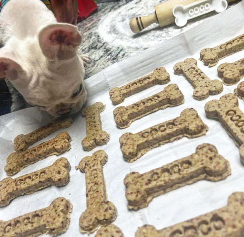 May include: Bone-shaped dog treats with the word "coco" imprinted on them are arranged on a white surface. A white and tan French Bulldog is looking down at the treats. A wooden rolling pin with a bone-shaped handle is in the background.