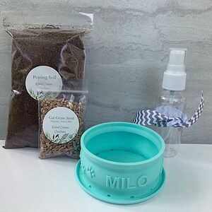 Peut inclure: Un pot &agrave; herbe &agrave; chat bleu clair avec le nom "MILO" sur le devant. Le pot est entour&eacute; d'un sac de terreau, d'un sac de graines d'herbe &agrave; chat et d'un flacon pulv&eacute;risateur transparent avec un bouchon blanc.