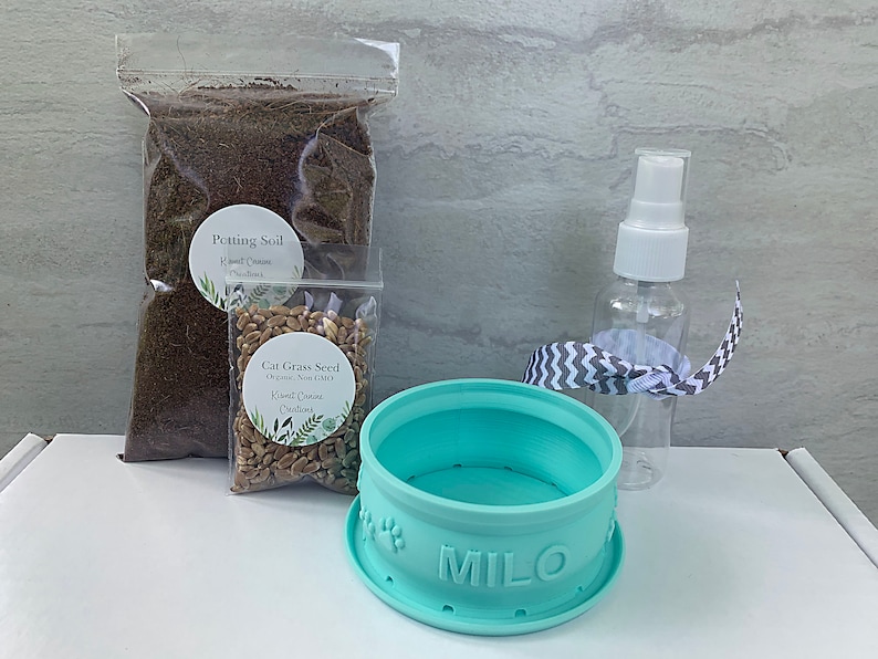 Peut inclure: Un pot &agrave; herbe &agrave; chat bleu clair avec le nom "MILO" sur le devant. Le pot est entour&eacute; d'un sac de terreau, d'un sac de graines d'herbe &agrave; chat et d'un flacon pulv&eacute;risateur transparent avec un bouchon blanc.