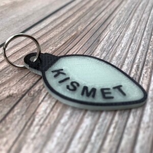 Op de afbeelding: Een witte lampvormige sleutelhanger met een zwarte omtrek en het woord "KISMET" in het zwart gedrukt.